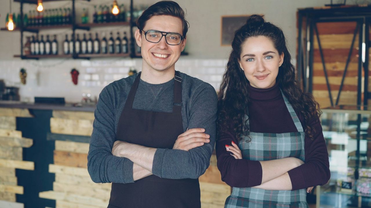 Two cafe owners in aprons, smiling with arms crossed behind their counter